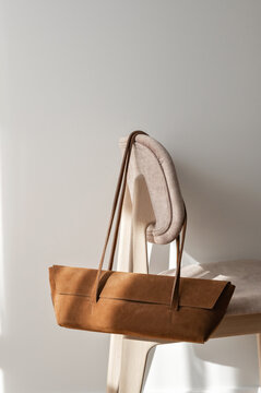 Caramel brown sueded shoulder baguette bag hanging on chair, neutral beige empty wall background with natural sunlight. Fashion woman accessory and clothing concept for business branding
