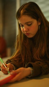 A focused young student is engrossed in writing or drawing with a green pen. The concentrated girl dedicates herself to her academic or creative work.