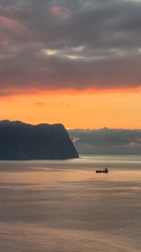 Fishing boat sails across calm ocean near coastal cliffs at golden hour sunrise. Static wide shot.