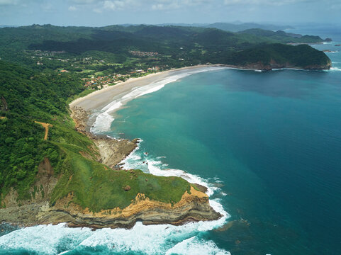 Aerial view of Playa La Redonda with its sandy beach, lush green cliffs, and turquoise ocean waves crashing against the rocky coastline in Tola, Rivas, Nicaragua.