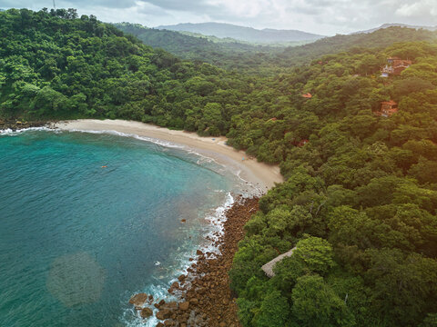 Aerial view of Playa La Redonda featuring a secluded sandy beach, turquoise waves crashing against rocky shores, and dense tropical forest with hillside villas in Tola, Rivas, Nicaragua.