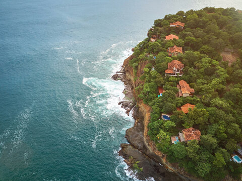 Aerial view of Playa La Redonda featuring coastal villas nestled in lush vegetation along the rocky shoreline with ocean waves crashing against the cliffs in Tola, Rivas, Nicaragua.