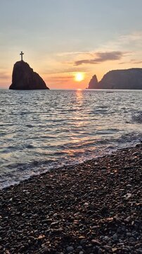 Sunset, sea, cross, beautiful sky over a serene rocky beach with ocean waves and distant mountains, peaceful twilight glow. Vertical video