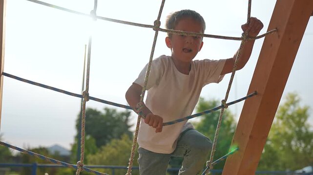 Boy climbing rope on playground. Boy plays actively on wooden structure. Boy enjoying outdoor adventure, developing strength. Fun playground activity, boy pulling rope. Happy childhood outdoor play.