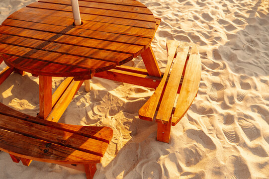 round wooden picnic table with attached benches resting on sunlit sand beside calm beach shoreline in warm