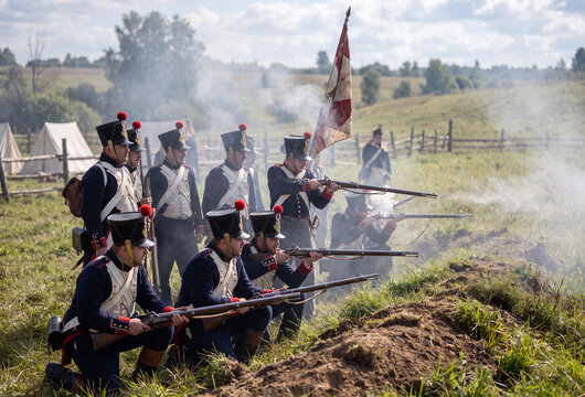 Napoleonic soldiers fire muskets during a reenactment on a grassy field with smoke billowing from their weapons.