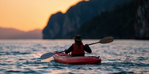Fototapeta premium Woman kayaking at sunset in red kayak with cliffs in the distance for outdoor adventure wallpaper and travel background design