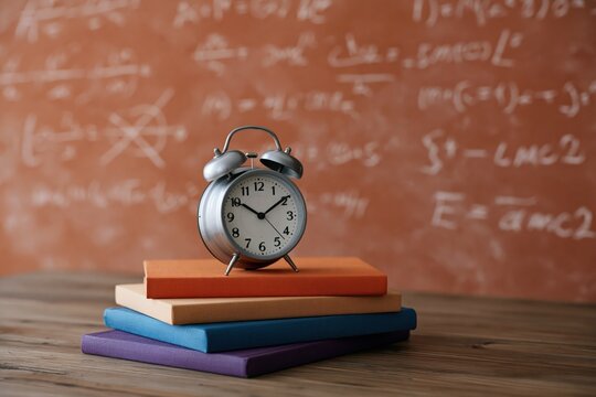 Silver alarm clock balanced on colorful book stack with blurred chalk equations for study and back to school background or banner design