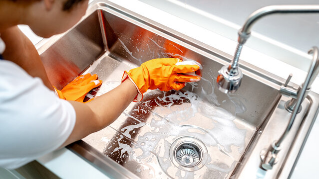 Asian male housekeeper with orange rubber glove scrubbing on kitchen sink surface with soap suds spreading around. Cleaning stainless steel sink for hygiene. Housework chores in the kitchen