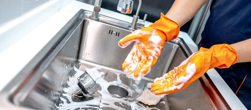 Housekeeper showing hands with orange rubber glove full with soap suds while scrubbing on kitchen sink surface. Cleaning stainless steel sink for hygiene. Housework chores in the kitchen