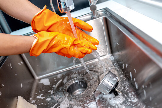 Housekeeper washing hands while wearing orange rubber gloves after cleaning the stainless steel kitchen sink.