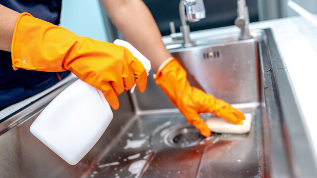 Housekeeper hands in orange rubber gloves holding detergent spray bottle while scrubbing stainless steel kitchen sink with sponge. Home hygiene maintenance service