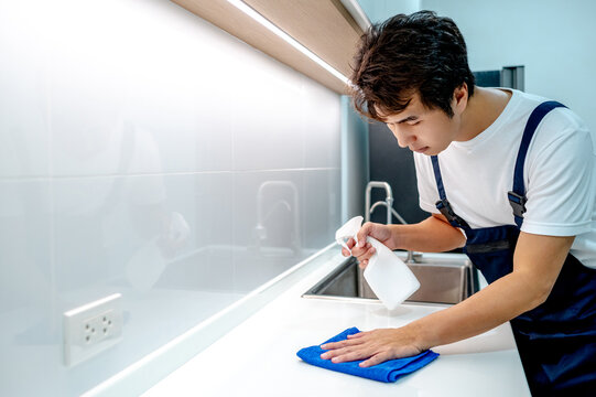Asian male housekeeper holding detergent spray bottle while wiping blue microfiber cloth on modern white countertop in domestic kitchen. Home hygiene maintenance. Cleaning house service
