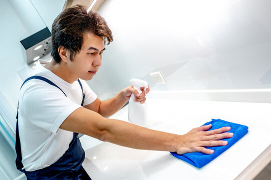 Asian male housekeeper holding detergent spray bottle while wiping blue microfiber cloth on modern white countertop in domestic kitchen. Home hygiene maintenance. Cleaning house service
