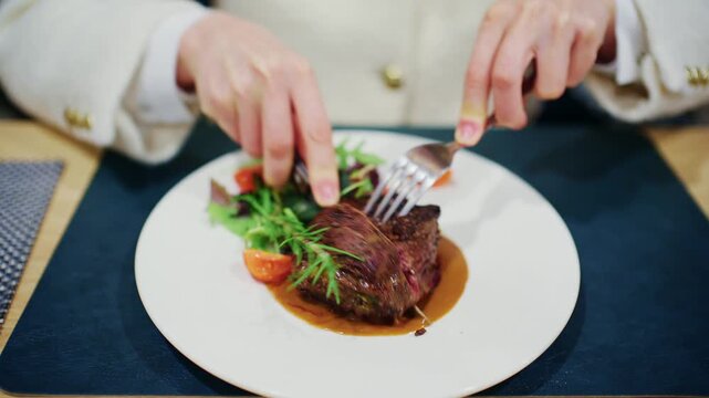 Person slicing a cooked steak with a knife and fork on a plate with salad in a restaurant setting.