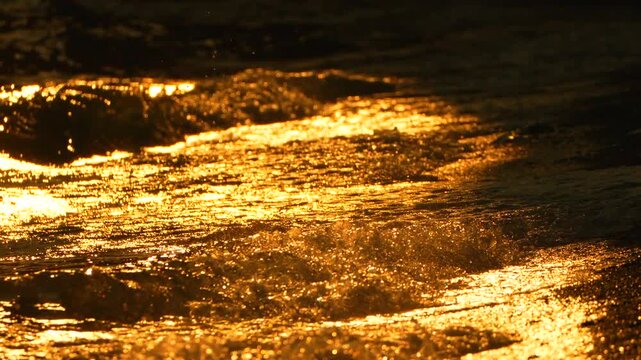 Glowing ocean waves crash against the sandy shoreline at sunset, creating brilliant orange light reflections. Static close-up shot.