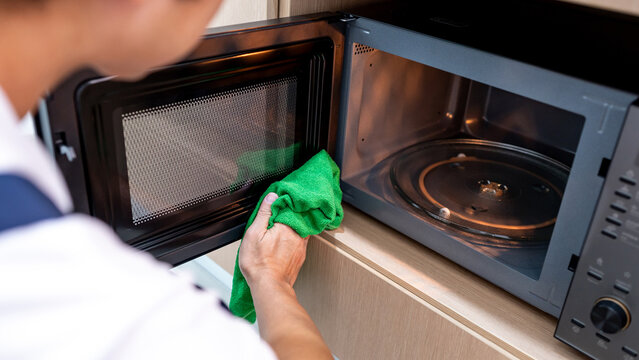 Male hand holding green microfiber cloth wiping inside the black microwave oven in wooden cabinet. Cleaning home appliance in the kitchen