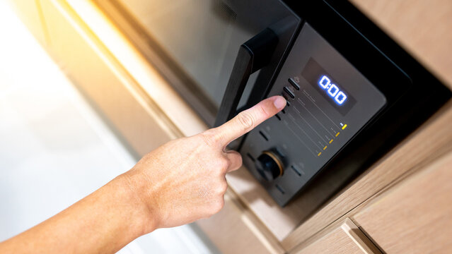 Hand pressing button setting mode and timer on modern black microwave oven in wooden cabinet. Using home appliance in the kitchen