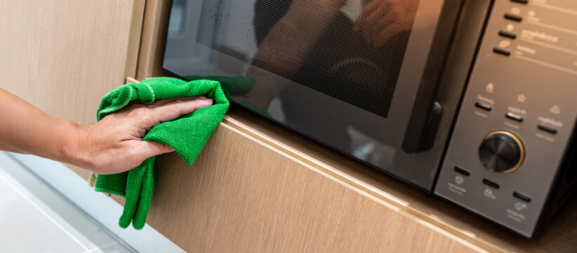 Male hand holding green microfiber cloth wiping on modern black microwave oven in wooden cabinet. Cleaning home appliance in the kitchen