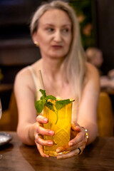 Fototapeta premium Woman extends a glass of non alcoholic mojito with lime, mint and ice toward the camera in a restaurant. Invitation to refreshment concept supporting hospitality, celebration and customer experience.