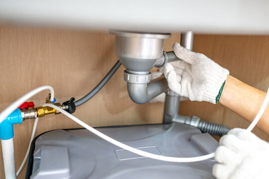 Plumber hand with glove fixing pipe connection and checking grease trap box under kitchen sink. Handyman repairing plumbing system in wooden cabinet.
