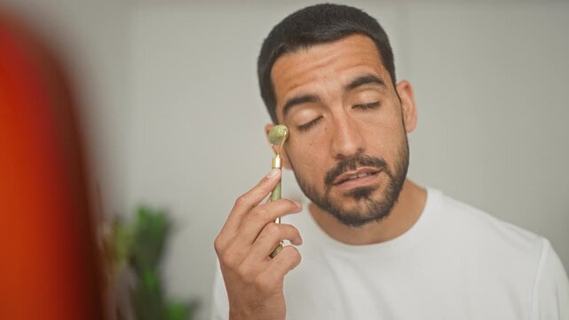 Man rolling jade facial roller on cheek in studio, eyes closed and holding the roller near jawline with a gentle gesture; wellness calm.