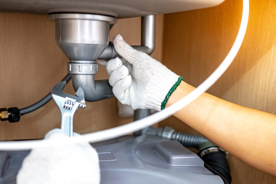Plumber hand with glove fixing pipe connection with adjustable wrench and checking grease trap box under kitchen sink. Handyman repairing plumbing system in wooden cabinet.