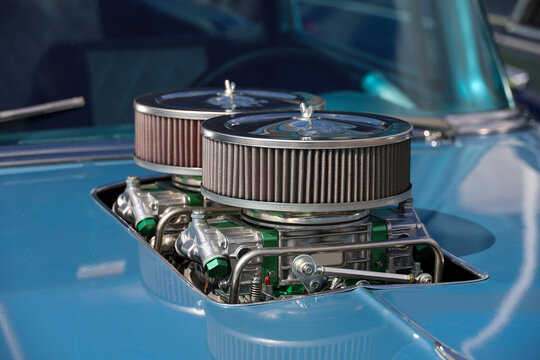 Close-up of a classic vintage car engine compartment featuring a dual-quad carburetor setup with custom chrome air cleaners protruding through a cut-out in the blue hood