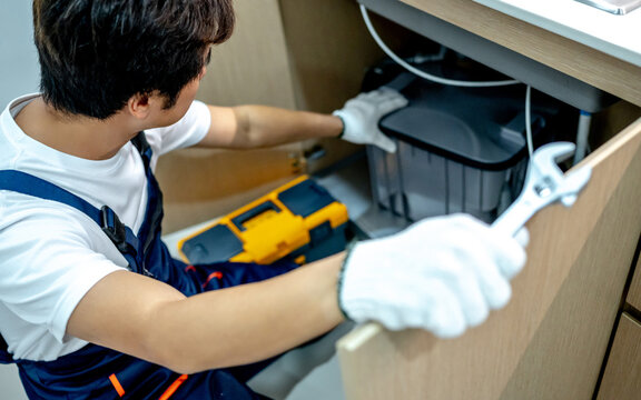 Asian male plumber with glove and adjustable wrench in his hand opening wooden cabinet checking grease trap box and pipe system under the kitchen sink. Handyman fixing plumbing system in the kitchen.