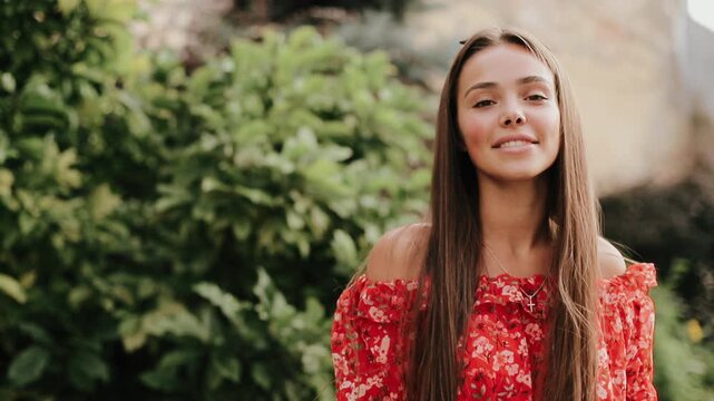 Young woman turns head from left toward right while smiling warmly and keeping relaxed posture in sunlit garden, with dense green shrubs and soft natural daylight behind, with young, woman, teen