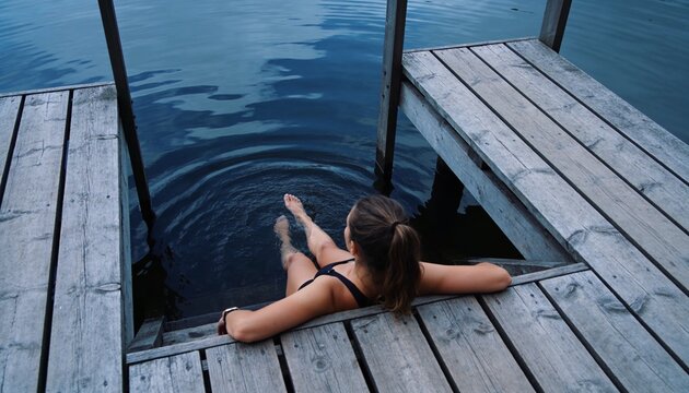 Reclining woman wearing black swimsuit and ponytail at lakeside wood dock with wristwatch, ripples