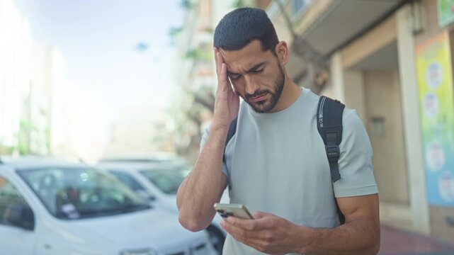 Young man holding smartphone with hand to temple on a sunlit street, backpack on shoulder and squinting at screen; stress urgency.