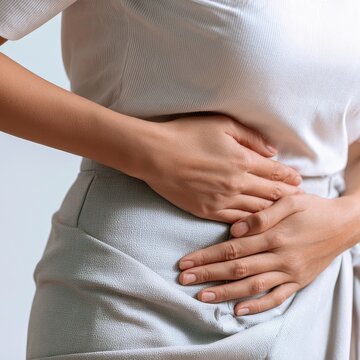 Female individual in a white shirt holds her stomach with both hands, displaying a concerned expression, against a light background emphasizing the subject's discomfort