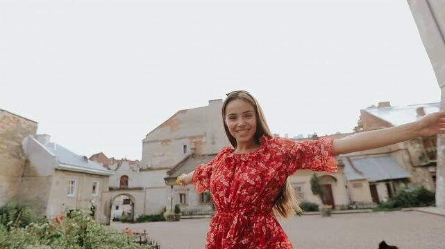 Young woman in red floral dress raises arms and holds small yellow flower while standing in historic town square, with stone buildings, soft overcast daylight, and cheerful travel mood