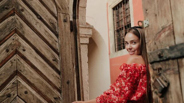 Young woman wearing red floral off shoulder dress opens weathered wooden gate beside rustic stone entrance, smiling back over shoulder with playful inviting expression and relaxed pose