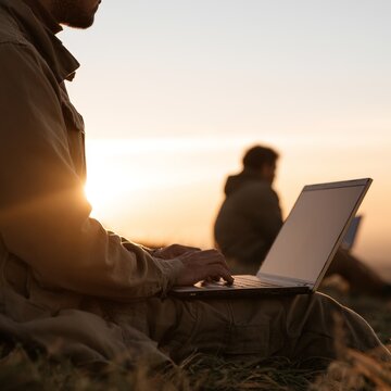 Male individual in casual attire using a laptop while seated on grass during sunset, with a blurred figure in the background and warm sunlight illuminating the scene