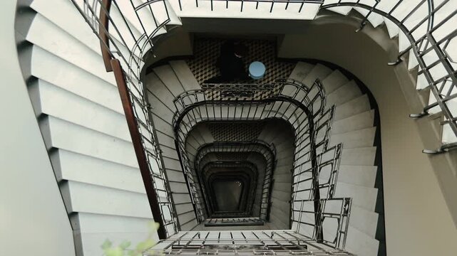 Overhead perspective looking straight down into deep indoor rectangular spiral stairwell, showing repeated concrete steps, glossy metal balustrades, wooden handrail accent, neutral tones, and