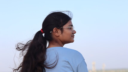 Side profile portrait of a thoughtful young Indian woman with wind-blown hair wearing glasses and a casual t-shirt, looking away towards the horizon against a clear sky background.  © Lovely