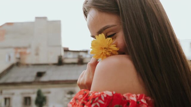 Young woman in red patterned dress holds yellow daisy near face, turning slightly with soft smile in bright courtyard, creating intimate, peaceful mood with gentle movement and warm daylight