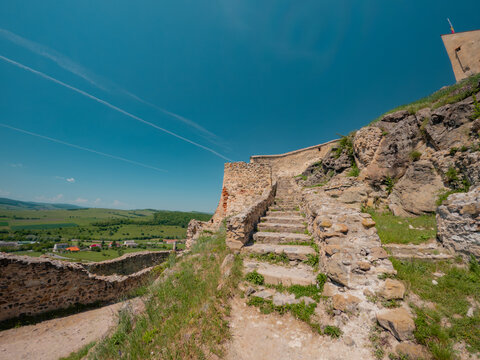Wide angle view framed by historic stone walls of Rupea Fortress, looking over the town below and green surrounding hills under a bright blue summer sky in Transylvania, Romania.