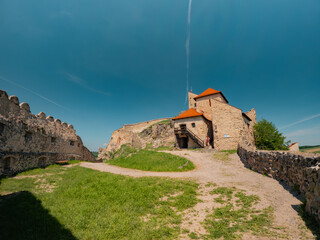 Wide angle view framed by historic stone walls of Rupea Fortress, looking over the town below and green surrounding hills under a bright blue summer sky in Transylvania, Romania. © badescu
