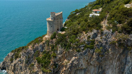 Naklejka premium Aerial view of a historical stone watchtower standing proudly on a high rocky cliff, overlooking vibrant turquoise sea waters and surrounded by lush green Mediterranean hillsides in a sunny morning.
