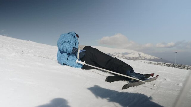 A person wearing ski gear sits on a pristine white snow-covered mountain. They are trying to stand up with their skis on a sunny winter day.
