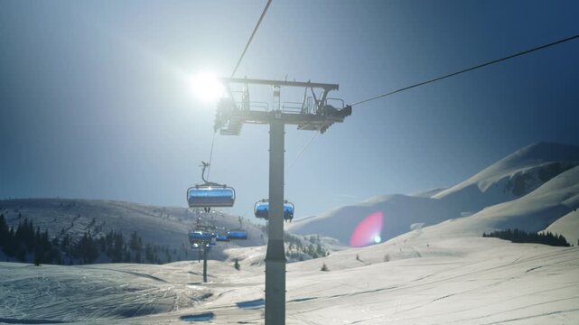 A modern chairlift ascends a snowy mountain slope with a clear blue sky overhead. Empty ski lift chairs move up the mountain on a bright winter day.