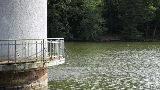 Metal railing and stone base of an old water tower on a lake in Germany, close-up of the weathered white wall and platform above the green water with a wooded shore behind, static shot