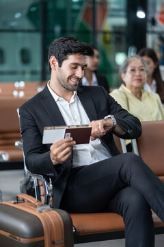young male executive in suit holding passport with boarding pass, sitting in modern airport terminal checking time on wrist watch,preparing for corporate business trip or international flight