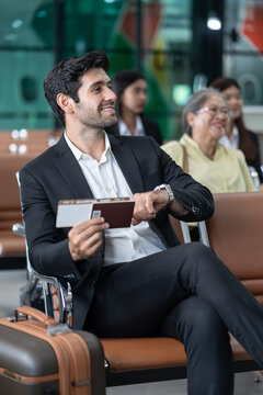 young male executive in suit holding passport with boarding pass, sitting in modern airport terminal checking time on wrist watch,preparing for corporate business trip or international flight