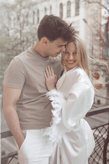 Fototapeta premium A girl in a white coat and a guy in a T-shirt and white pants are standing on a balcony against the background of a city street. A love story