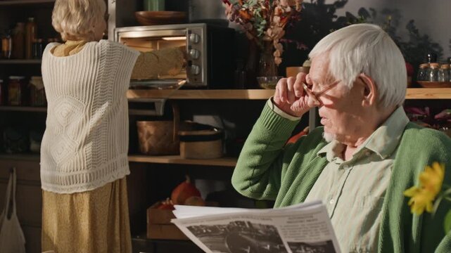 Senior Caucasian man reading newspaper while senior Caucasian woman brings freshly baked pie to table, zooming out. Clip is ideal for retirement lifestyle and senior care marketing campaigns