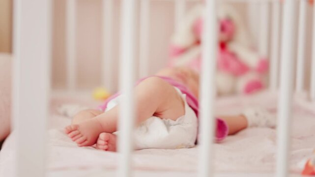 Little baby in white diaper lying on soft mattress inside nursery crib.